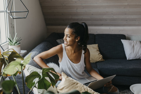 Junge Frau sitzt mit Laptop auf der Couch und schaut aus dem Fenster, lizenzfreies Stockfoto