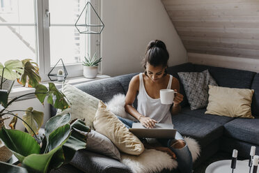 Young woman sitting on the couch with cup of coffee using laptop - LHPF00009