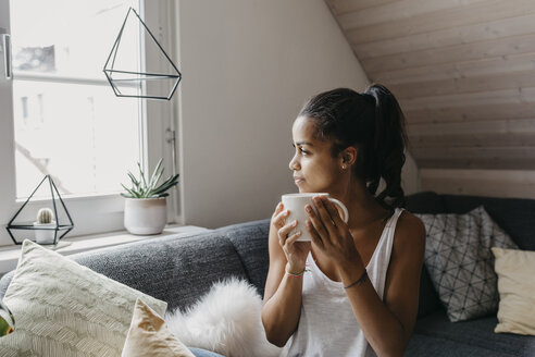 Junge Frau mit einer Tasse Kaffee, die zu Hause auf der Couch sitzt und aus dem Fenster schaut - LHPF00010