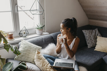 Young woman with laptop and cup of coffee sitting on the couch at home looking out of window - LHPF00011