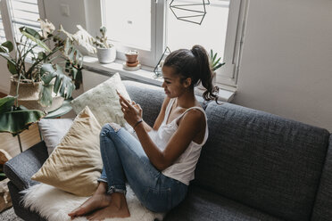 Young woman sitting on the couch at home using mobile phone - LHPF00015
