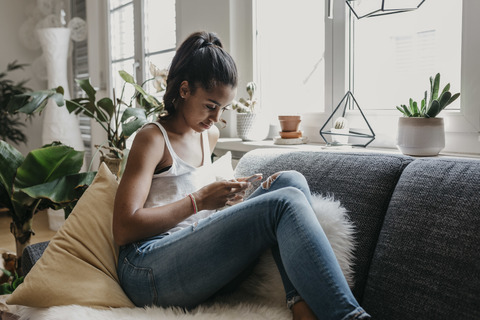 Junge Frau sitzt zu Hause auf der Couch und benutzt ein Mobiltelefon, lizenzfreies Stockfoto