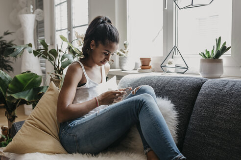 Young woman sitting on the couch at home using mobile phone - LHPF00017