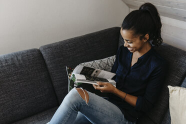 Junge Frau sitzt auf der Couch zu Hause und liest eine Zeitschrift - LHPF00023