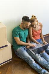 Ein Paar sitzt nebeneinander auf dem Boden eines neuen Hauses und schaut gemeinsam auf einen Laptop - FBAF00018