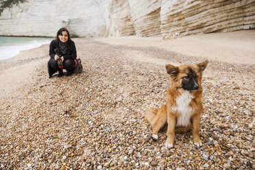 Italy, Vieste, stray dog sitting on Vignanotica Beach while smiling woman crouching in the background - FLMF00008