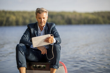 Porträt eines entspannten Geschäftsmannes mit Kopfhörern und Tablet auf einem Paddelbrett am See sitzend - FMKF05209