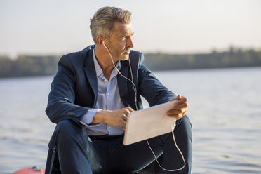 Mature businessman with earphones and tablet relaxing at lake - FMKF05210