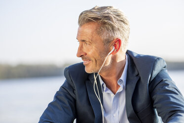 Smiling businessman listening music with earphones at lake - FMKF05212