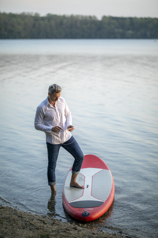 Älterer Mann mit Kopfhörern und Tablet am Seeufer stehend mit Paddleboard, lizenzfreies Stockfoto