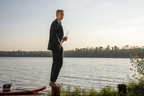 Geschäftsmann mit Getränk steht barfuß auf einer Stange am See, lizenzfreies Stockfoto