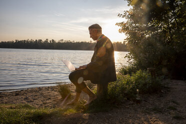 Mature businessman sitting at lakeshore by sunset working on laptop - FMKF05235