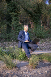 Mature businessman with laptop sitting barefoot in nature looking at distance - FMKF05236