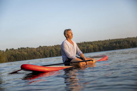 Älterer Mann sitzt auf Paddleboard auf einem See bei Sonnenuntergang entspannen, lizenzfreies Stockfoto