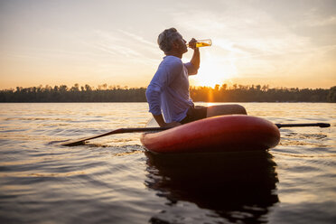 Man sitting on paddleboard on a lake by sunset drinking beverage - FMKF05240