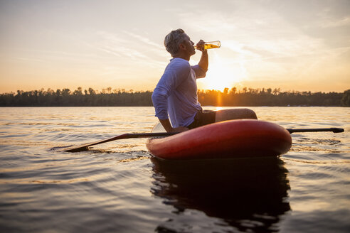 Man sitting on paddleboard on a lake by sunset drinking beverage - FMKF05240