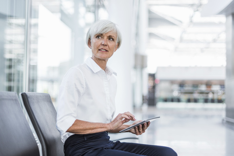 Ältere Geschäftsfrau sitzt im Wartebereich mit Tablet und schaut sich um, lizenzfreies Stockfoto