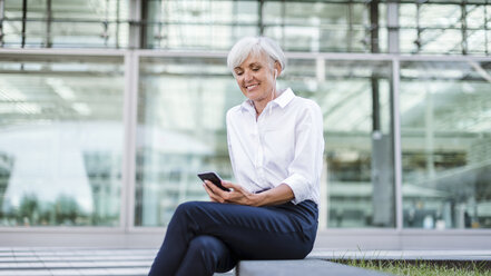 Ältere Geschäftsfrau sitzt draußen mit Smartphone und Ohrstöpseln - DIGF05038