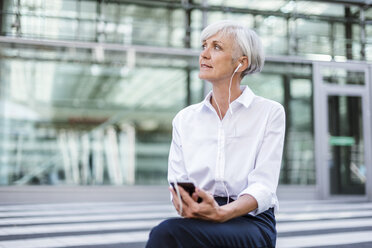 Ältere Geschäftsfrau sitzt draußen mit Smartphone und Ohrstöpseln - DIGF05039