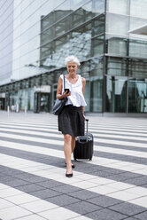 Smiling senior woman with baggage on the move looking on cell phone - DIGF05088