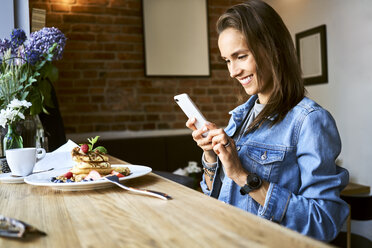 Smiling young woman with plate of pancakes using phone in cafe - BSZF00576