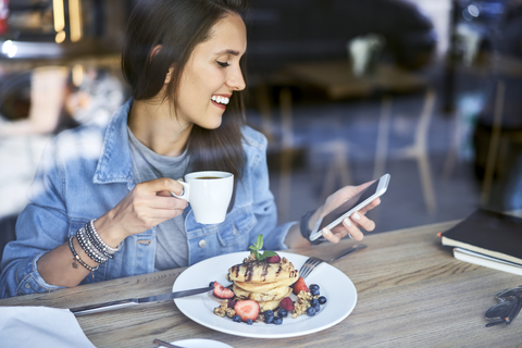 Lächelnde junge Frau mit einem Teller Pfannkuchen, die in einem Café telefoniert, lizenzfreies Stockfoto