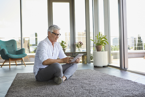 Mature man sitting on carpet at home using tablet stock photo