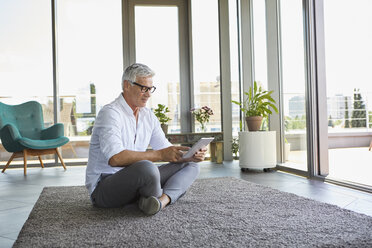 Älterer Mann sitzt zu Hause auf einem Teppich und benutzt ein Tablet - RBF06528