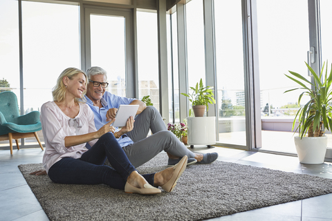 Smiling mature couple relaxing at home sitting on carpet sharing tablet stock photo