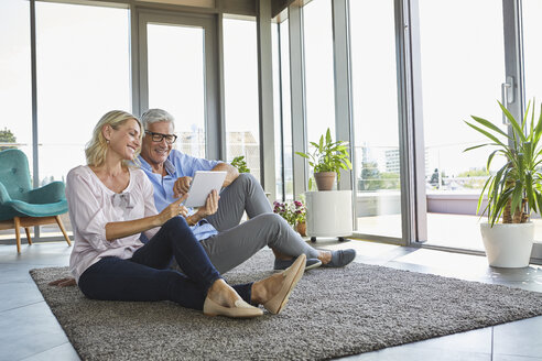 Smiling mature couple relaxing at home sitting on carpet sharing tablet - RBF06552