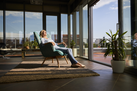 Reife Frau entspannt im Sessel im Sonnenlicht zu Hause, lizenzfreies Stockfoto