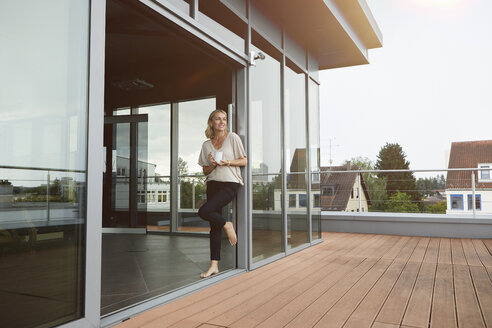 Smiling mature woman with cup of coffee standing at roof terrace at home - RBF06574