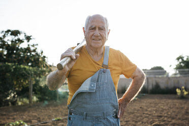 Senior man in denim overall holding garden tool on shoulder and looking at camera - JRFF01834