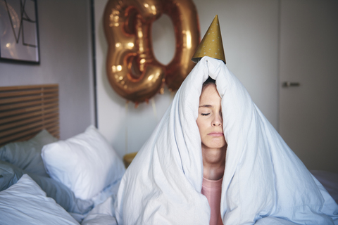 Sad woman celebrating her birthday, sitting on bed under blanket stock photo