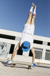 Rückenansicht eines Mannes in stilvollem, sportlichem Outfit, der auf einem Skateboard steht, kopfüber vor blauem Himmel - JRFF01844