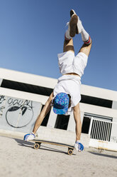 Rückenansicht eines Mannes in stilvollem, sportlichem Outfit, der auf einem Skateboard steht, kopfüber vor blauem Himmel - JRFF01845