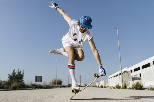 Mann in stilvollem sportlichem Outfit steht auf einem Skateboard vor blauem Himmel - JRFF01850