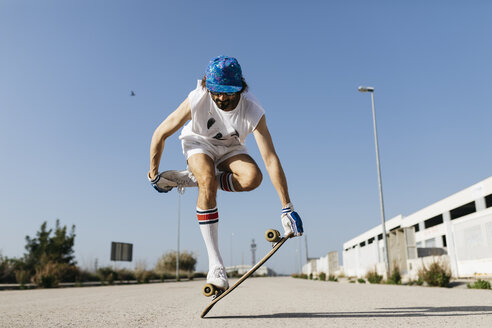 Mann in stilvollem sportlichem Outfit steht auf einem Skateboard vor blauem Himmel - JRFF01851