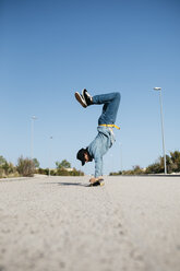 Trendy man in denim and cap skateboarding, standing on skateboard upside down - JRFF01864