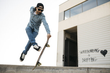Trendy man in denim and cap skateboarding, doing jump with skateboard from concrete ramp - JRFF01866