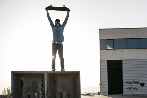 Skateboarder in Freizeitkleidung auf einer Holzkiste mit dem Skateboard über dem Kopf, lizenzfreies Stockfoto