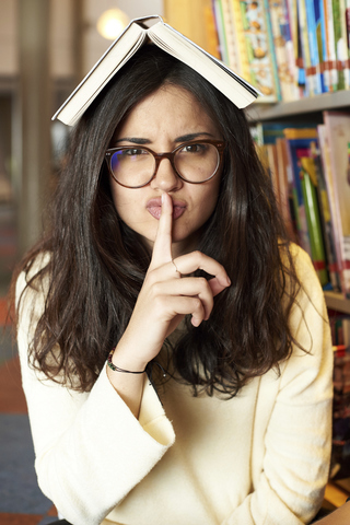Portrait of young woman with book on top of head asking for silence at the library stock photo