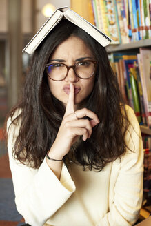 Porträt einer jungen Frau mit einem Buch auf dem Kopf, die in der Bibliothek um Ruhe bittet - IGGF00570