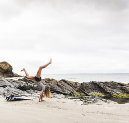 Junge Frau mit Surfbrett macht Handstand am Strand - UUF15029