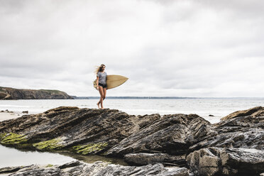 Young woman carrying surfboard on a rocky beach at the sea - UUF15033