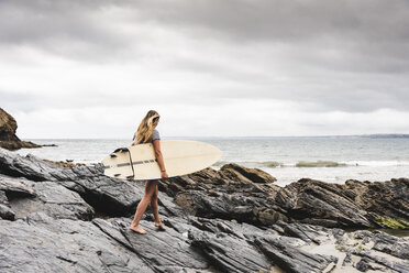 Junge Frau mit Surfbrett an einem felsigen Strand am Meer - UUF15036