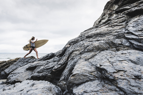 Junge Frau mit Surfbrett an einem felsigen Strand am Meer, lizenzfreies Stockfoto