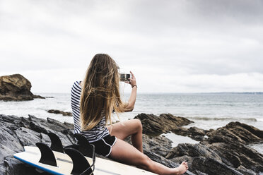 Junge Frau mit Surfbrett am Strand sitzend, mit Smartphone - UUF15040