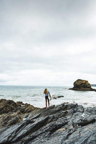 Junge Frau mit Surfbrett an einem felsigen Strand am Meer, lizenzfreies Stockfoto