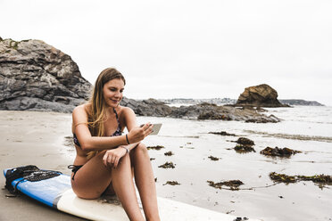 Junge Frau mit Surfbrett am Strand sitzend, mit Smartphone - UUF15048